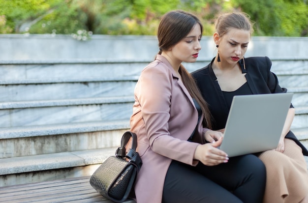 Dwie kobiety biznesu siedzą na ławce z laptopem i patrzą na ekran na zewnątrz