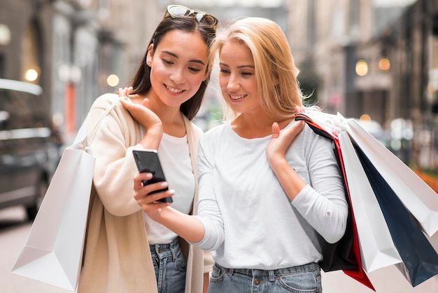 Dwie kobiety na zewnątrz patrząc na smartfona, trzymając torby na zakupy