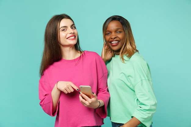 Dwie kobiety przyjaciele europejski i afroamerykanin w różowe zielone ubrania trzymać w ręku telefon komórkowy na białym tle na tle niebieskiej ściany turkus, portret studio. Koncepcja życia ludzi. Makieta miejsca na kopię.