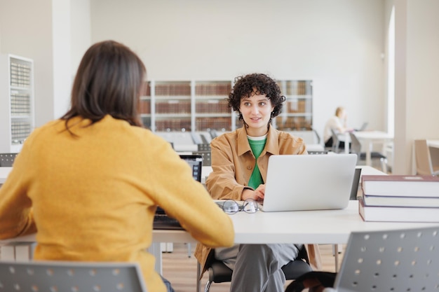 Dwie kobiety w bibliotece lub coworkingu siedzą naprzeciwko siebie i pracują na laptopie