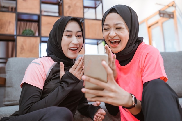 Dwie Kobiety W Hidżabie śmieją Się Podczas Wspólnego Czytania Wiadomości Przez Telefon Komórkowy, Siedząc Na Podłodze W Domu