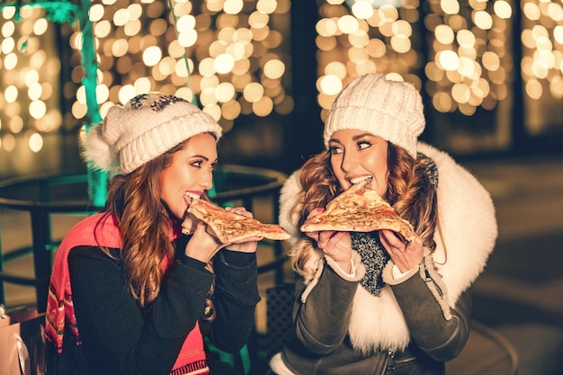 Dwie koleżanki spędzają wieczór na mieście, patrząc na siebie i śmiejąc się z kawałkami pizzy w dłoniach.