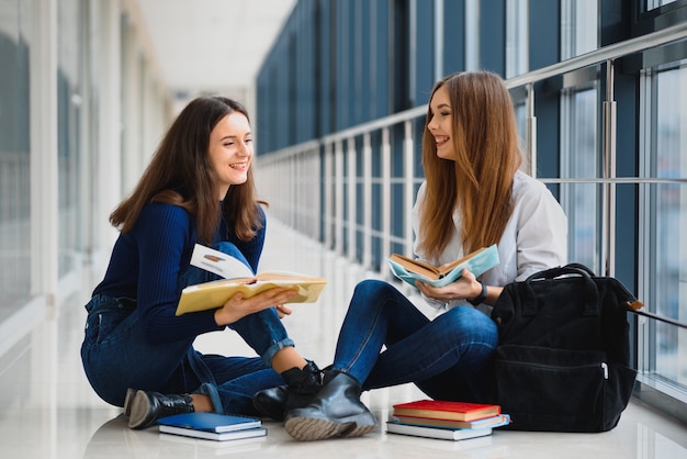 Dwie ładne studentki z książkami siedzą na podłodze w korytarzu uniwersytetu