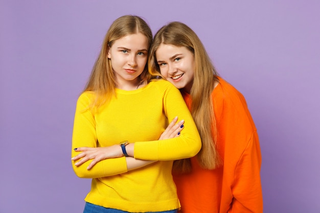 Dwie ładne uśmiechnięte młode blondynki bliźniaczki siostry dziewczyny w żywych kolorowych ubraniach stojących, odizolowane na pastelowej fioletowej niebieskiej ścianie. Koncepcja życia rodzinnego osób.