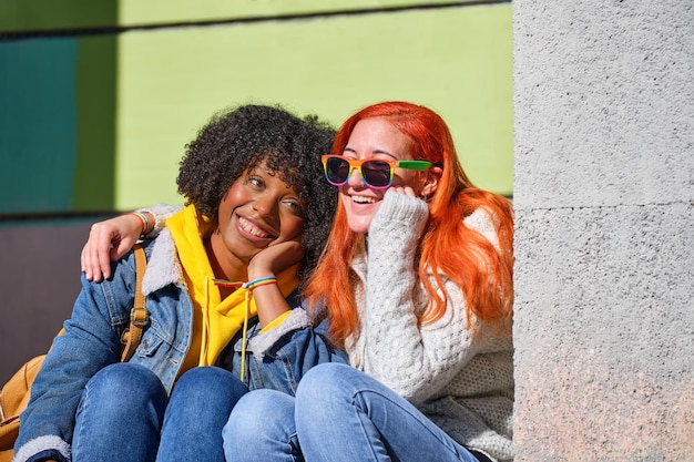 Dwie lesbijki siedzą, obejmując jedną białą z pomarańczowymi włosami i drugą czarną z włosami afro
