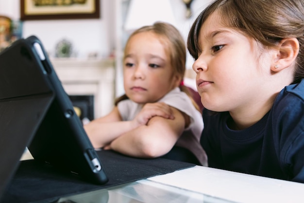 Dwie Małe Dziewczynki Uważnie Oglądają Wideo Na Tablecie W Swoim Salonie Koncepcja Uczenia Się Technologii W Dzieciństwie, Zabawy Z Internetem I łącznością