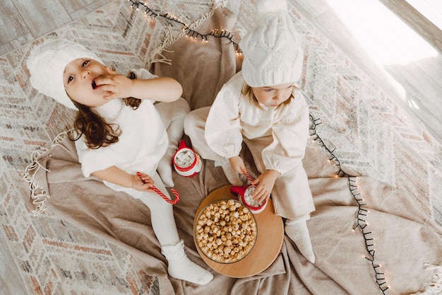 Dwie małe dziewczynki w czapkach zimowych siedzą na podłodze w pobliżu choinki i jedzą popcorn. koncepcja bożego narodzenia.