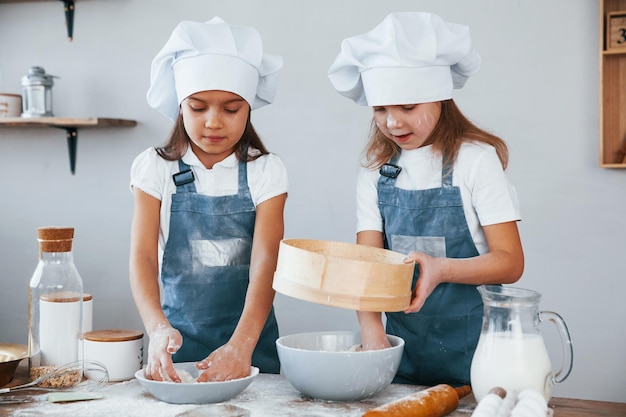 Dwie małe dziewczynki w niebieskim mundurze szefa kuchni pracujące z mąką za pomocą sita w kuchni