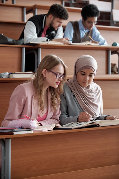 Dwie międzykulturowe studentki college'u lub uniwersytetu siedzące w pierwszym rzędzie przy biurku w sali wykładowej i omawiające fragment w książce na lekcji