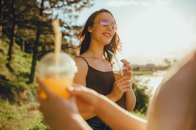 Dwie miłe koleżanki, bawiące się latem, pijące sok w okularach przeciwsłonecznych, w biało-czarnej koszuli, o przyrodzie, o zachodzie słońca, pozytywny wyraz twarzy, outdoor