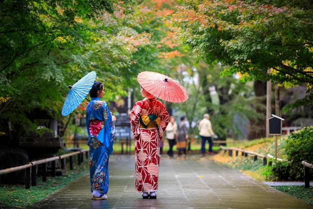 Dwie młode azjatyckie kobiety w niebieskim i czerwonym kimono z parasolem na ręce spacerują po parku w sezonie jesiennych liści w japonii