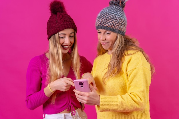 Dwie młode blondynki rasy kaukaskiej z telefonem odizolowanym na różowym tle uśmiechniętym z internetem