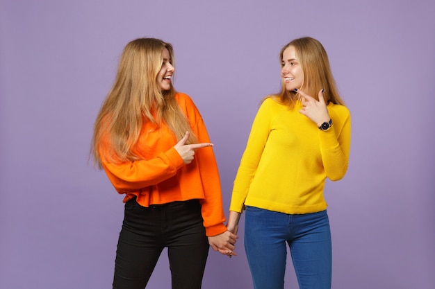 Dwie młode blondynki siostry bliźniaczki dziewczyny w żywych ubraniach, wskazując palcem wskazującym na siebie na białym tle na pastelowej fioletowej niebieskiej ścianie. Koncepcja życia rodzinnego osób.
