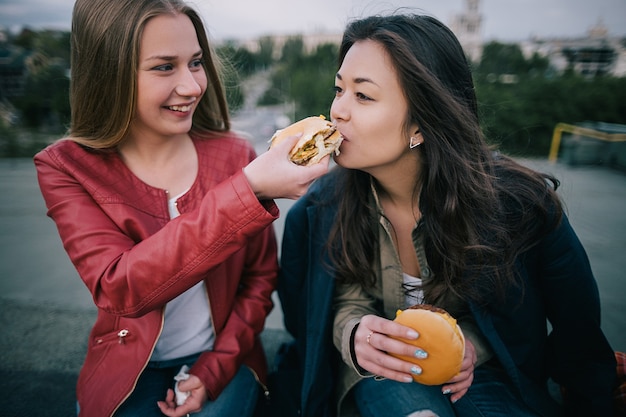 Zdjęcie dwie młode dziewczyny dzielące się smacznym burgerem z bliska. wspólne spędzanie czasu na dachu, niezwykłe miejsca wypoczynku i rozrywki, wypoczynek w wesołej atmosferze with