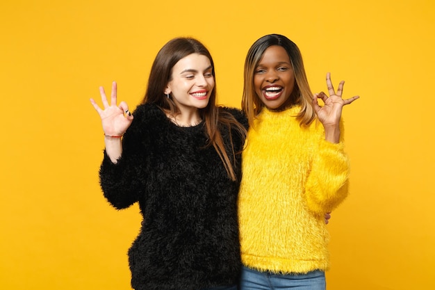 Dwie młode kobiety przyjaciele europejski i afroamerykanin w czarne żółte ubrania stojący pozowanie na białym tle na tle jasnej pomarańczowej ściany, portret studio. Koncepcja życia ludzi. Makieta miejsca na kopię.