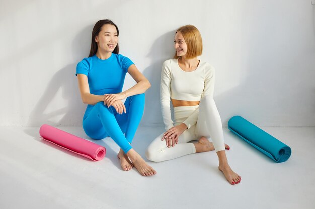Dwie młode kobiety siedzące na zajęciach jogi uśmiechają się i rozmawiają