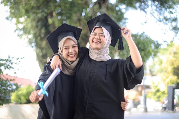 Dwie młode muzułmańskie damy w strojach dyplomowych pozujące przed kamerą na kampusie uniwersyteckim z dyplomem