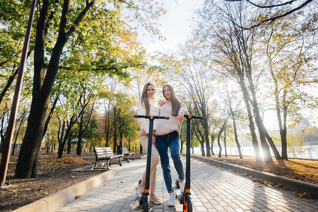 Dwie młode piękne dziewczyny jeżdżą na skuterach elektrycznych w parku w ciepły jesienny dzień.
