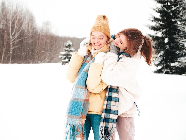Dwie młode piękne uśmiechnięte hipster kobieta w modne ciepłe ubrania i szaliki. Beztroskie kobiety pozowanie na ulicy w parku. Pozytywne modele czystej zabawy na śniegu. Ciesz się zimowymi chwilami. Świąteczna koncepcja