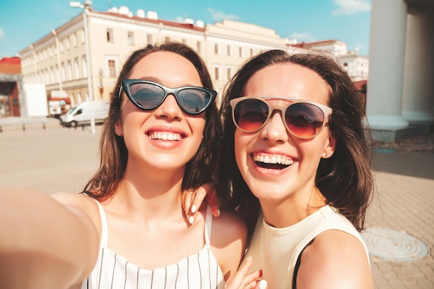 Dwie młode piękne uśmiechnięte kobiety hipster w modnych letnich ubraniachSeksowne beztroskie kobiety pozują na tle ulicy w kapeluszu Pozytywne czyste modele bawią się o zachodzie słońca robiąc zdjęcia Pov w mieście