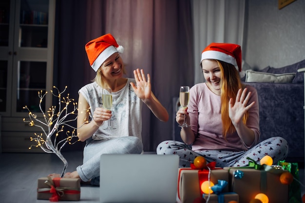 Dwie młode radosne kobiety w czerwonych czapkach Świętego Mikołaja, siedzące na podłodze, korzystające z laptopa i komunikujące się zdalnie z przyjaciółmi i rodziną. Noworoczne życzenia z szampanem w przytulnym wnętrzu domu.