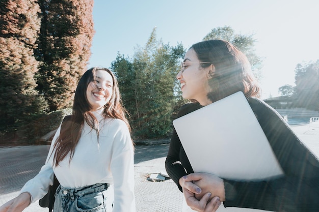 Dwie młode studentki idzie do klasy, rozmawiając i śmiejąc się na zewnątrz wydziału w jasny dzień. Studia uniwersyteckie, papiery studenckie, styl nowoczesny. Przygotowanie prezentacji na zajęcia.