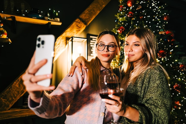 Dwie Młode Szczęśliwe Kobiety Robiące Selfie świąteczne Zdjęcie W Pobliżu Choinki I Picia Wina.