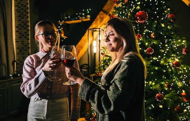 Zdjęcie dwie młode szczęśliwe kobiety wznoszące toast i pijące wino w pobliżu choinki.