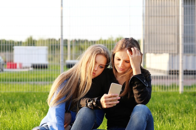 Dwie nastoletnie dziewczęta używające smartfona przy selfie siedzące razem Dwie dziewczyny śmiejące się i grimacing