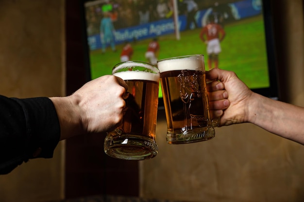 Dwie osoby wznoszące toast szklankami piwa przed telewizorem z telewizorem pokazującym mecz piłki nożnej na ekranie