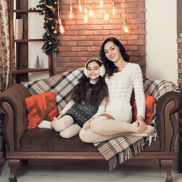 Dwie piękne dziewczyny, matka i córka siedzące na kanapie w świątecznie urządzonym pokoju.