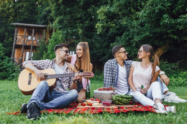 Dwie piękne dziewczyny z dwoma chłopcami siedzą w parku na kocu z gitarą, mają piknik i słuchają melodii gitary