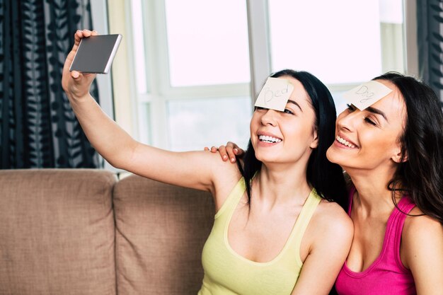 Dwie piękne i zabawne dziewczyny z wklejonymi kartkami papieru na czołach grają w grę i robią sobie zdjęcia lub robią selfie