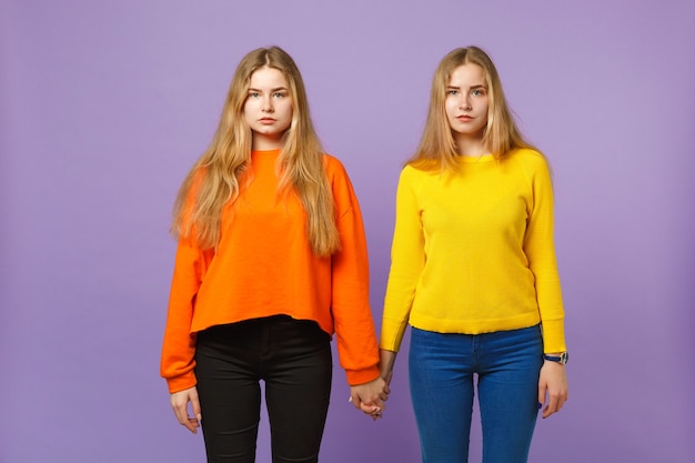 Dwie piękne młode blond siostry bliźniaczki dziewczyny w żywe kolorowe ubrania stojące, na białym tle na pastelowej fioletowej niebieskiej ścianie. Koncepcja życia rodzinnego osób.