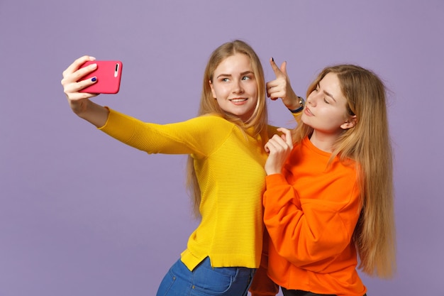 Dwie piękne młode blond siostry bliźniaczki dziewczyny w żywych ubraniach robi selfie nakręcony na telefon komórkowy na białym tle na pastelowej fioletowej niebieskiej ścianie. Koncepcja życia rodzinnego osób.