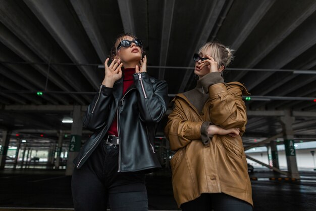Dwie piękne młode dziewczyny w stylu młodzieżowym ze skórzaną kurtką i czarnymi dżinsami z okularami przeciwsłonecznymi pozują na parkingu na ulicy