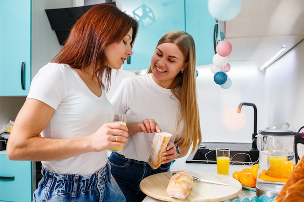 Dwie piękne młode koleżanki stoją w domu przy kuchennym stole i jedzą chleb i piją sok pomarańczowy