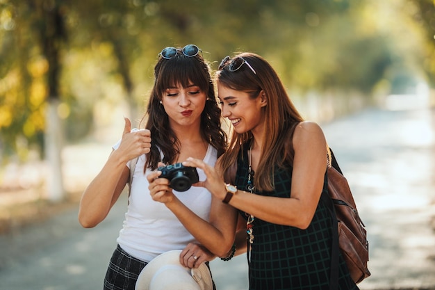 Dwie piękne młode uśmiechnięte kobiety z plecakami na plecach patrzą na zdjęcia w aparacie podczas spaceru po jesiennej słonecznej alei.