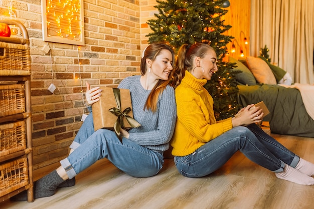 Dwie piękne wesołe szczęśliwe dziewczyny młode dziewczyny dają prezenty świąteczne na tle drzewa nowego roku w domu