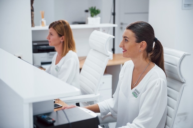 Dwie pielęgniarki w białym mundurze siedzą w recepcji szpitala i wykonują swoją pracę.