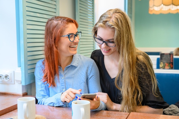 Dwie podekscytowane młode dziewczyny korzystające z telefonów komórkowych siedzące w kawiarni i wskazujące palcem Rudowłosa kobieta w okularach pokazuje swojej przyjaciółce śmieszne zdjęcia na smartfonie dziewczyna blondynka Piją kawę
