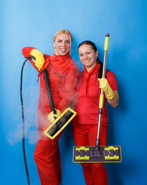 Dwie pracownice firmy sprzątającej w czerwonych mundurach uśmiechają się na odosobnionym niebieskim tle