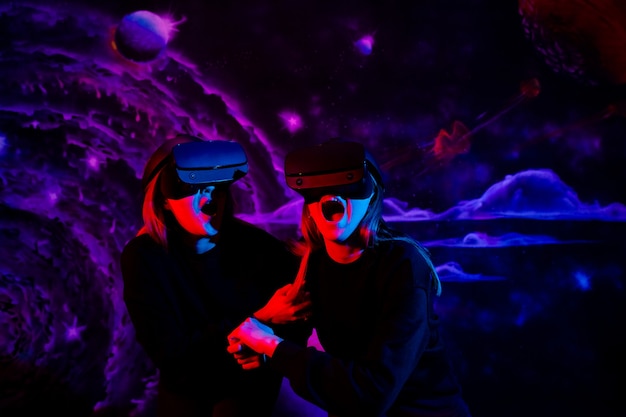Dwie przyjaciółki siostry dziewczyny z wirtualnymi okularami trzymającymi się za ręce bawią się w pokoju gier w neonowym niebieskim i czerwonym świetle