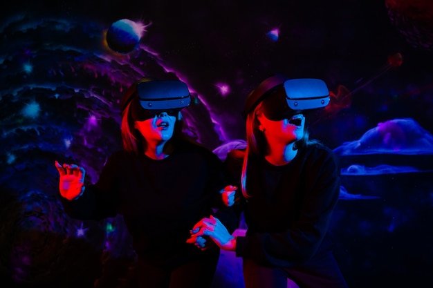 Dwie przyjaciółki w wirtualnych okularach trzymają się za ręce w pokoju zabaw w neonowym świetle