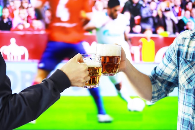 Dwie ręce poziome trzymając szklankę piwa lager i brzęk na tle meczu piłki nożnej. Fani sportu rozweselają. Koncepcja stylu życia wypoczynku przyjaciół.