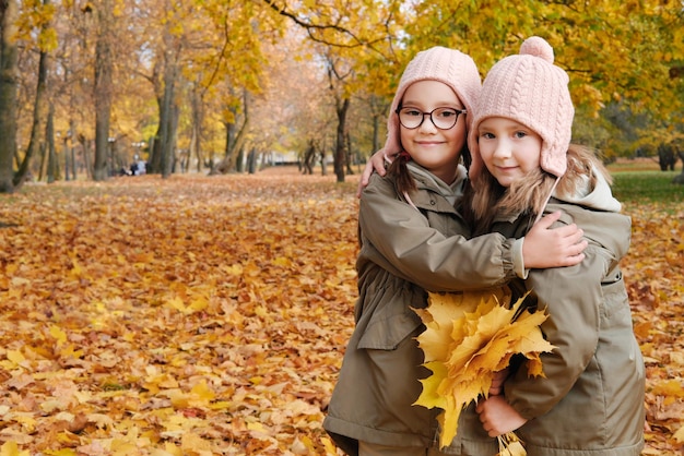 Dwie siostry bliźniaczki pozują do kamery z bukietem żółtych liści w jesiennym parku