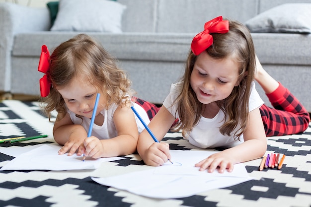 Dwie Siostry Dziewczynki Leżą Na Podłodze Domu I Rysują Kredkami Na Papierze.