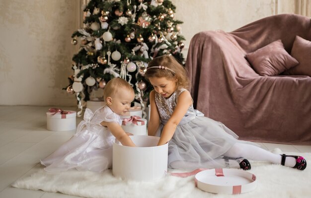Dwie śliczne dziewczynki w świątecznych sukienkach siedzą na kocu obok choinki i otwierają pudełko