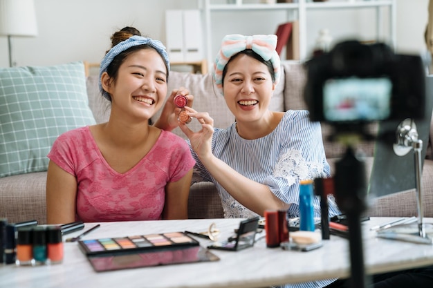 Dwie śliczne dziewczyny blogerki prezentujące kosmetyki kosmetyczne i transmitujące wideo na żywo w sieci społecznościowej. koncepcja vloga dla obserwujących nastolatków. młode kobiety patrzące w kamerę, śmiejące się, dobrze się bawiące