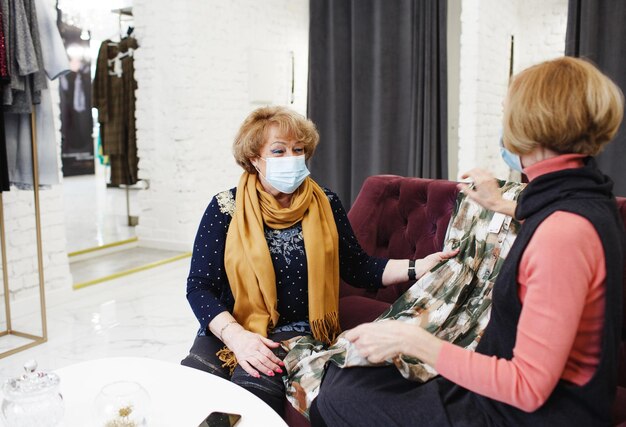 Dwie starsze kobiety w medycznych maskach patrzą na ubrania siedzące na kanapie w sklepie odzieżowym Zakupy i zakupy